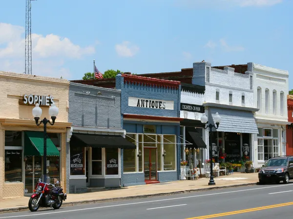 A row of brick and painted storefronts on Main Street in downtown Pineville, North Carolina, with an antiques shop, green and black awnings and old-style lamp posts under a blue sky
